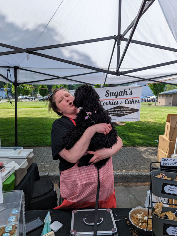 A woman stands behind a farmer's market stand, her wares of doggy cookies, cupcakes and other treats on display. She is holding the puppy and making an AWWWWWWWW face as the puppy gently kisses her cheek.