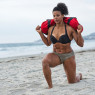 A dark-skinned woman on the beach in a dark bikini, doing lunges with a large sandbag across her shoulders.