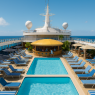 A bright expanse of blue sky & turquoise water surrounds the ship’s most lively escape.  Rows of loungers frame sparkling pools, palm-fringed cabanas offer shade, and the golden swim-up bar glows beneath the midday sun.