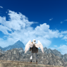 A screenshot of Clydwyn. He stands on a sloped stone surface in front of a gray mountain range with his back facing the viewer. He looks up high at a wide blue sky and its many clouds.